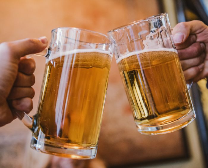 Twee glazen bier klinken op een proost, met schuimend bier dat uit de glazen spat en een achtergrond van een gezellige sfeer.